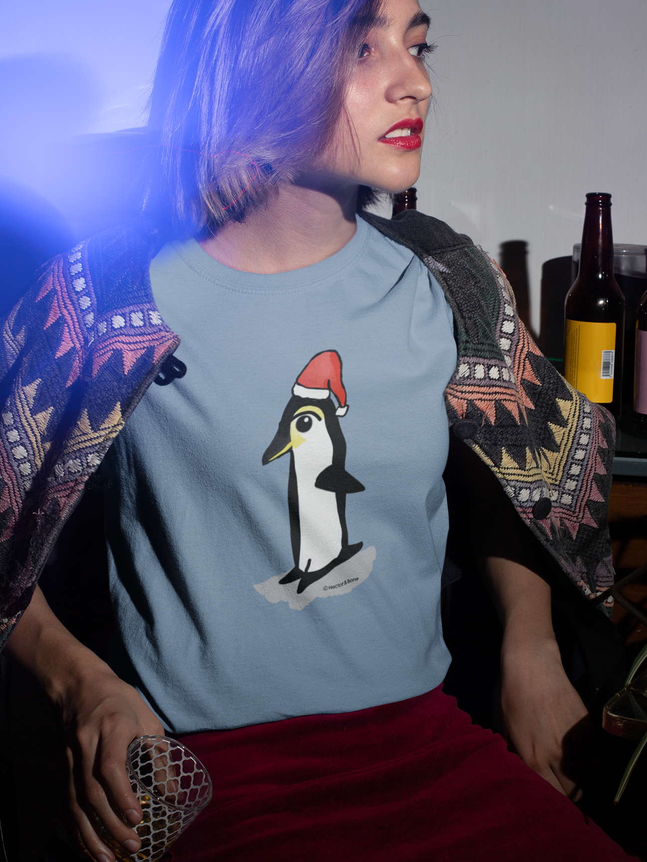 Christmas Penguin T-shirt - Young woman at a Christmas party wearing a Santa Penguin cute Christmas T-shirt in mid heather blue colour illustrated design by Hector and Bone on a Vegan cotton t-shirt